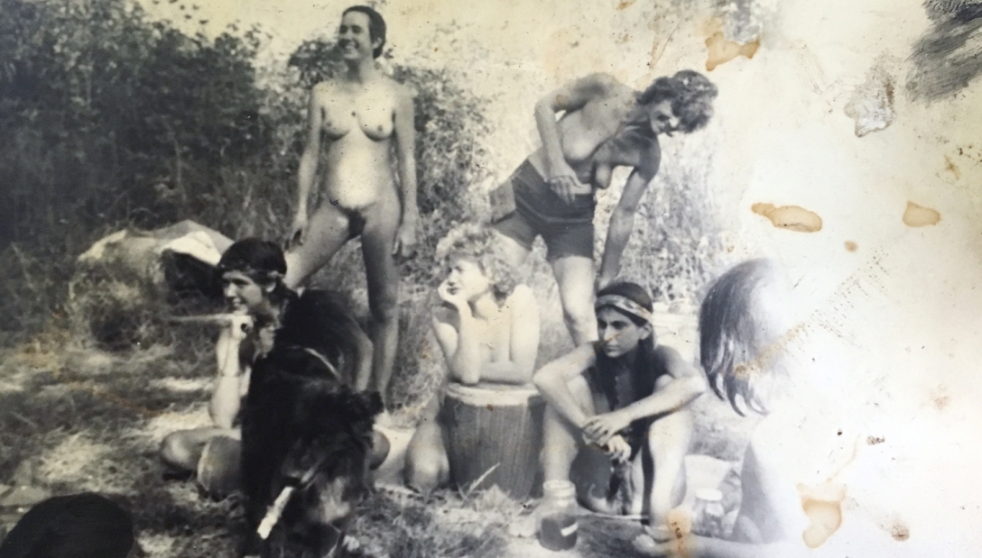 Foto de 6 mujeres en la comunidad rural separatista. Algunas están desnudas o parcialmente desnudas, entre árboles y junto a perros.